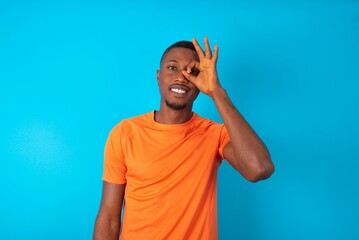Handsome man wearing orange T-shirt over blue background doing ok gesture with hand smiling, eye...