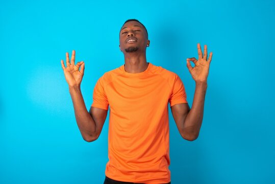 Handsome man wearing orange T-shirt over blue background relax and smiling with eyes closed doing meditation gesture with fingers. Yoga concept.