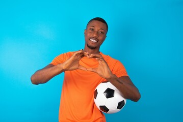 Man wearing orange T-shirt holding a ball over blue background smiling in love doing heart symbol...
