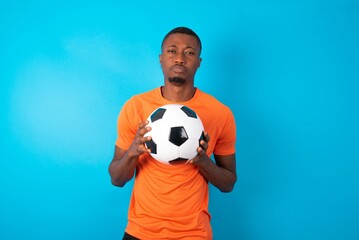 Displeased upset Man wearing orange T-shirt holding a ball over blue background frowns face as going to cry, being discontent and unhappy as can't achieve goals,  Disappointed model has troubles