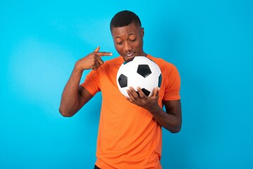 Shocked Man wearing orange T-shirt holding a ball over blue background look surprisedly down,...