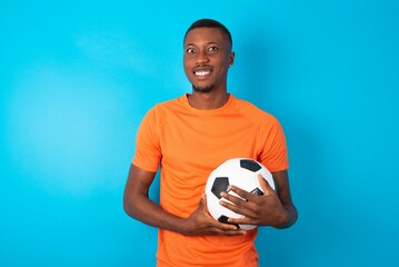 Man wearing orange T-shirt holding a ball over blue background keeps teeth clenched, frowns face in...