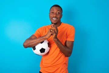 Positive Man wearing orange T-shirt holding a ball over blue background smiles happily, glad to receive pleasant news from interlocutor, keeps hands together. People emotions concept.