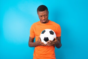 Dismal gloomy rejected Man wearing orange T-shirt holding a ball over blue background has problems...