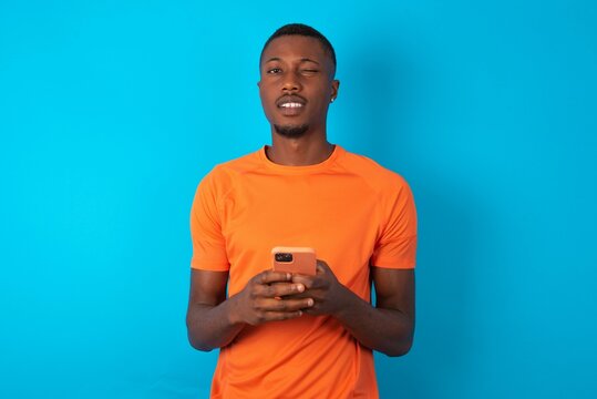 Handsome man wearing orange T-shirt over blue background taking a selfie  celebrating success