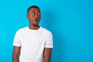 Shocked Handsome man wearing white T-shirt over blue background look empty space with open mouth...