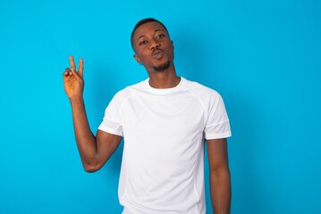 Handsome man wearing white T-shirt over blue background makes peace gesture keeps lips folded shows v sign. Body language concept