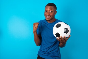 Young man wearing sport T-shirt holding a ball over blue background clenches fists and awaits for something nice happened looks away bites lips and waits announcement of results