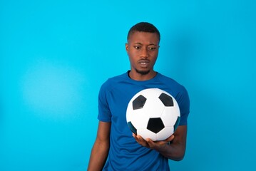 Portrait of dissatisfied Young man wearing sport T-shirt holding a ball over blue background smirks...