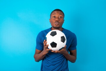 Young man wearing sport T-shirt holding a ball over blue background with surprised expression keeps hands under chin keeps lips folded makes funny grimace