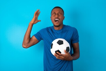 Young man wearing sport T-shirt holding a ball over blue background goes crazy as head goes around...