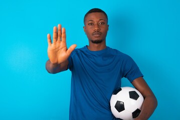 Young man wearing sport T-shirt holding a ball over blue background shows stop sign prohibition symbol keeps palm forward to camera with strict expression