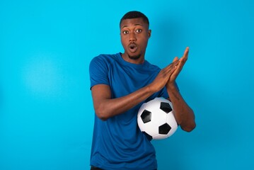 Surprised emotional Young man wearing sport T-shirt holding a ball over blue background rubs palms and stares at camera with disbelief