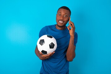 Smiling Young man wearing sport T-shirt holding a ball over blue background talks via cellphone, enjoys pleasant great conversation. People, technology, communication concept