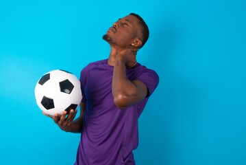 Man wearing purple T-shirt holding a ball over blue background suffering from back and neck ache injury, touching neck with hand, muscular pain.