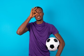 Playful excited Man wearing purple T-shirt holding a ball over blue background showing Ok sign with both hands on eyes, pretending to wear spectacles.