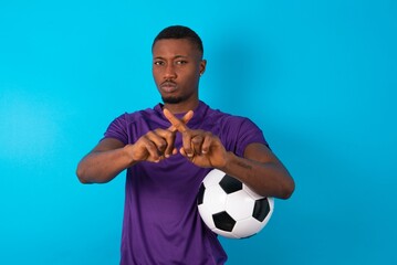 Man wearing purple T-shirt holding a ball over blue background has rejection angry expression...