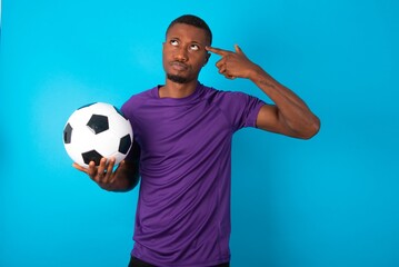 Unhappy Man wearing purple T-shirt holding a ball over blue background curves lips and makes...