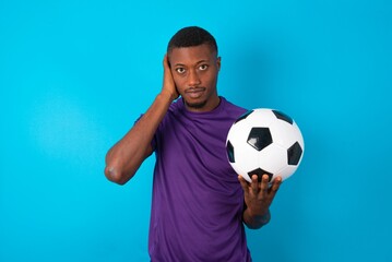 Frustrated Man wearing purple T-shirt holding a ball over blue background plugging ears with hands does not wanting to listen hard rock, noise or loud music.