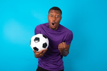 Joyful excited lucky Man wearing purple T-shirt holding a ball over blue  background cheering,...