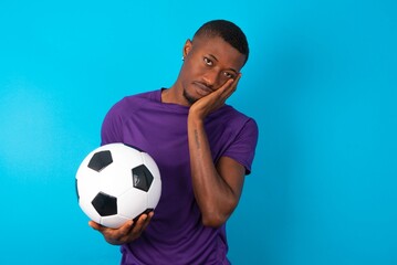 Very bored Man wearing purple T-shirt holding a ball over blue background holding hand on cheek while support it with another crossed hand, looking tired and sick.