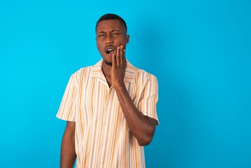 Tooth ache concept. young man wearing fashion shirt over blue background feeling pain, holding his cheek with hand, suffering from bad toothache, looking at camera with painful expression