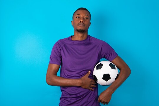 Picture of angry Man wearing purple T-shirt holding a ball over blue background looking camera.