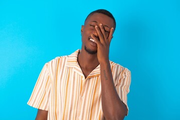 Handsome man wearing fashion shirt over blue background makes face palm and smiles broadly, giggles positively hears funny joke poses