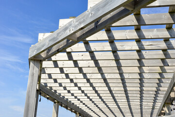 A grey colored worn and weathered wood pergola roof with a blue sky in the background. The outdoor sun shelter has wooden slats hanging over the wooden beams with string lights hanging off the boards.