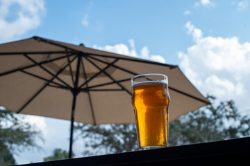 A clear pint beer glass filled with cold froth from a lager ale. The Irish red ale drink sits on the edge of a metal patio table at a craft microbrewery. There's a patio umbrella in the background.