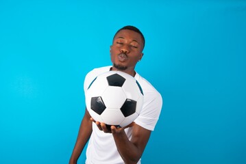 Young man wearing white T-shirt holding a ball over blue background looking at the camera blowing a kiss with hand on air being lovely. Love expression.
