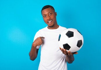 Young man wearing white T-shirt holding a ball over blue background being in stupor shocked, has astonished expression pointing at oneself with finger saying: Who me?