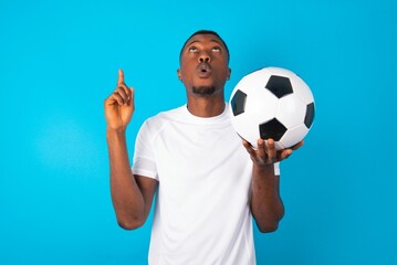 Young man wearing white T-shirt holding a ball over blue background being amazed and surprised...