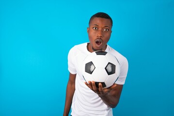 Young man wearing white T-shirt holding a ball over blue background having stunned and shocked...