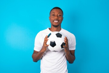 Young man wearing white T-shirt holding a ball over blue background keeps palms together, has...
