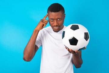 Young man wearing white T-shirt holding a ball over blue background concentrating hard on an idea with a serious look, thinking with both index fingers pointing to forehead.