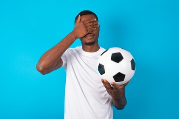Young man wearing white T-shirt holding a ball over blue background covering eyes with both hands,...