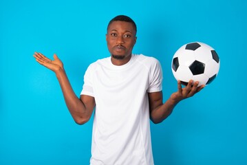 Puzzled and clueless Young man wearing white T-shirt holding a ball over blue background with arms out, shrugging shoulders, saying: who cares, so what, I don't know.