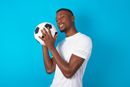 Dreamy Young man wearing white T-shirt holding a ball over blue background with pleasant expression, closes eyes, keeps hands crossed near face, thinks about something pleasant