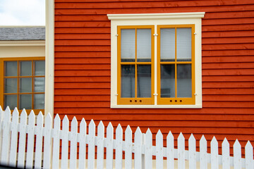 An exterior wall with orange-colored horizontal cape cod clapboard siding and a double-hung window. The closed vintage glass window has yellow and cream-colored trim with a white fence in front.