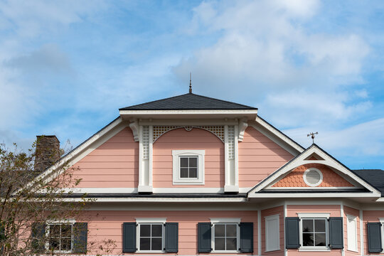 A multi story pink painted wooden house with white trim. The building has multiple small four pane glass windows with black shutters. There's a hip roof with black shingles and a metal weather vane. 