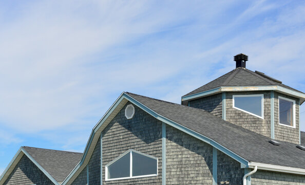A grey weathered wooden house with a rounds shaped turret. The large real estate property has small windows, white trim, and wooden siding on the building. The windows are unique geometric shapes. 