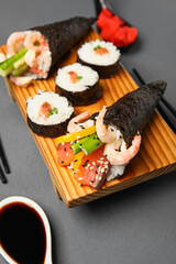 Wooden board with tasty sushi cones, rolls and soy sauce on dark background, closeup