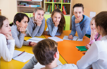 Friendly smiling female teacher talking to children, sitting together around desk in classroom