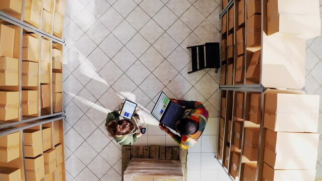 Top view of diverse depot employees working in storehouse, using tablet and laptop to check logistics and inventory list. Delivery team working on packing merchandise. Drone shot. Aerial view.