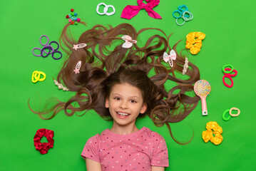 Hair accessories. A happy little girl lies surrounded by elastic bands and hair clips. Hairstyles for children. Green isolated background.