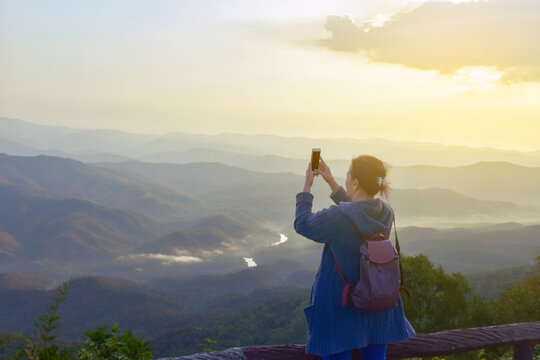 Asian elderly woman holding smartphone, standing near fence on the cliff and taking the photo of early morning view of mountain and landscapes, concept for happiness of elderly people after retirement
