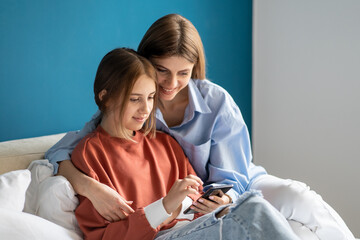 Positive mom daughter watching funny video in smartphone together sitting in bed at home. Family...