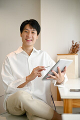 A handsome young Asian man sits in a minimal bright coffee shop with his digital tablet.