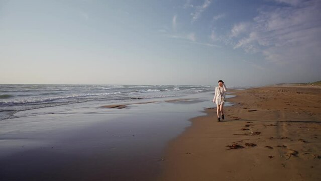 白いジャケット、黒いショートパンツの若い女性が夕日の浜辺を歩く風景 Young woman in white jacket and black shorts walking on beach at sunset 